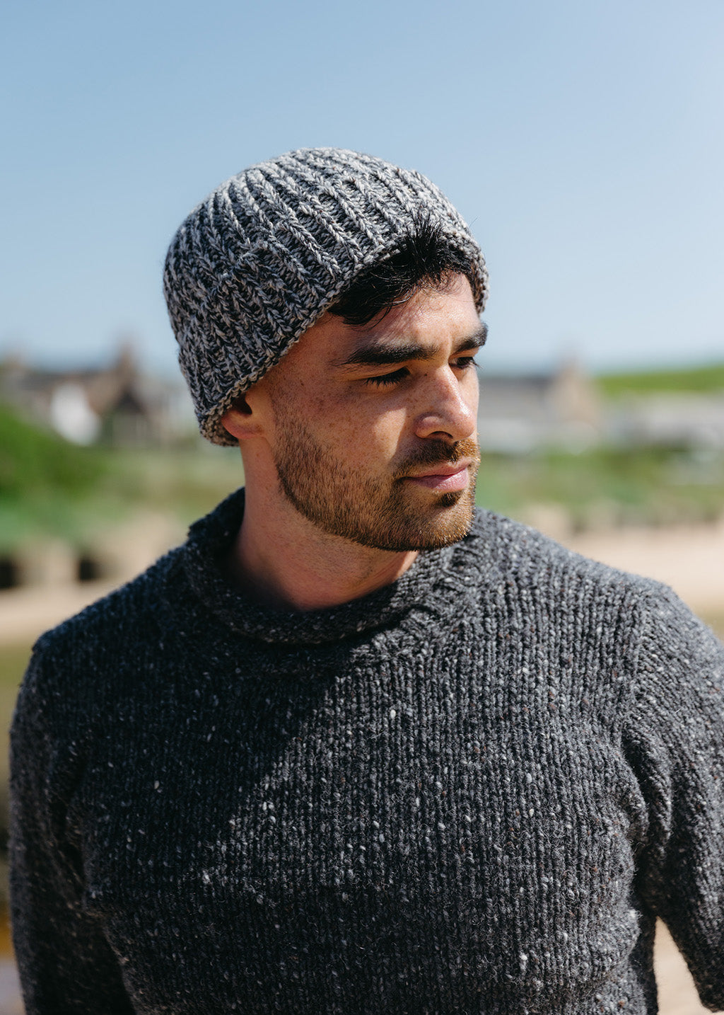 A man wears a mid grey marled chunky beanie hat with dark grey flecked jumper against an outdoor green blurred background.
