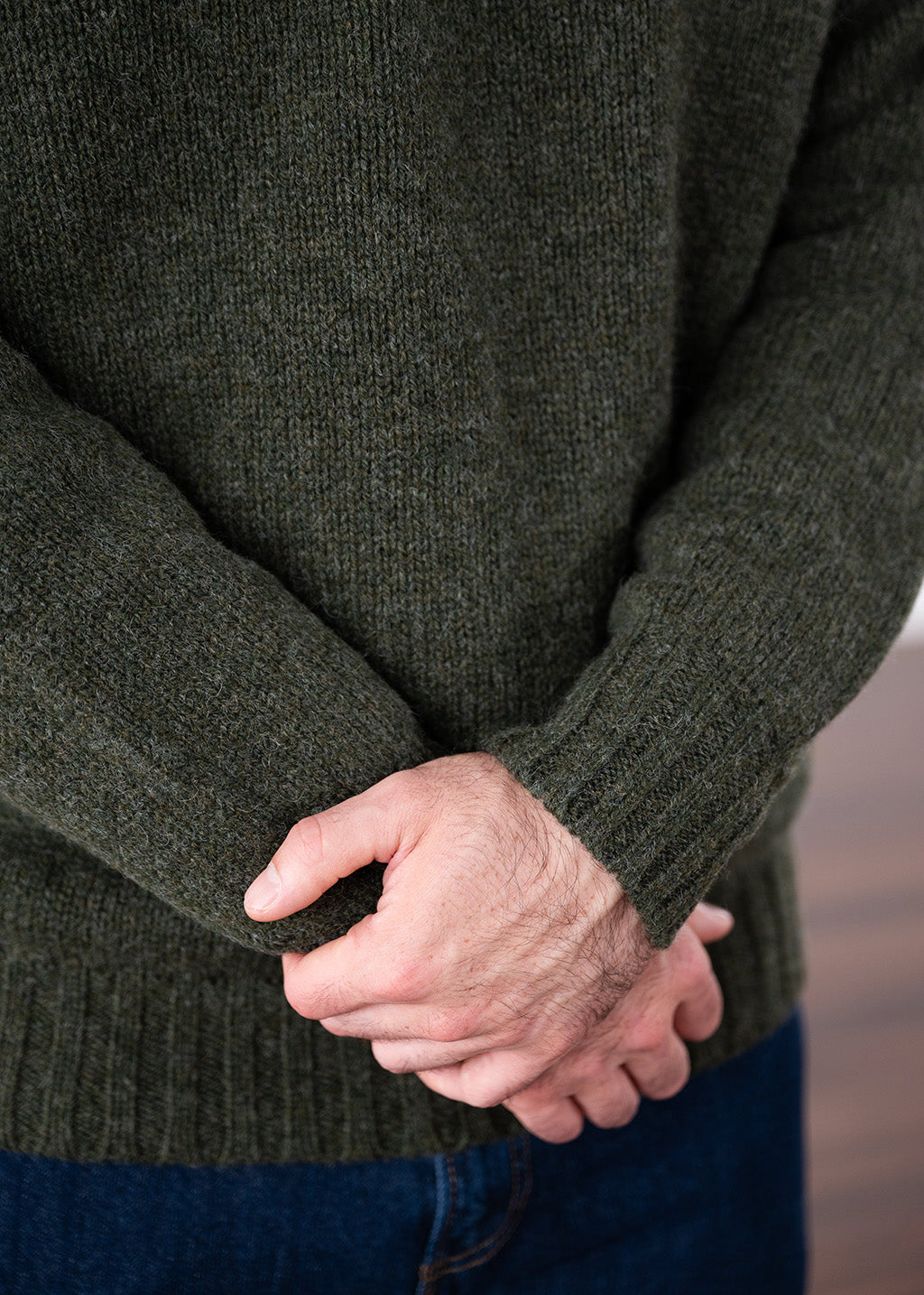 Close-up of a person wearing a dark olive green wool knit jumper, with hands clasped in front, paired with blue jeans.