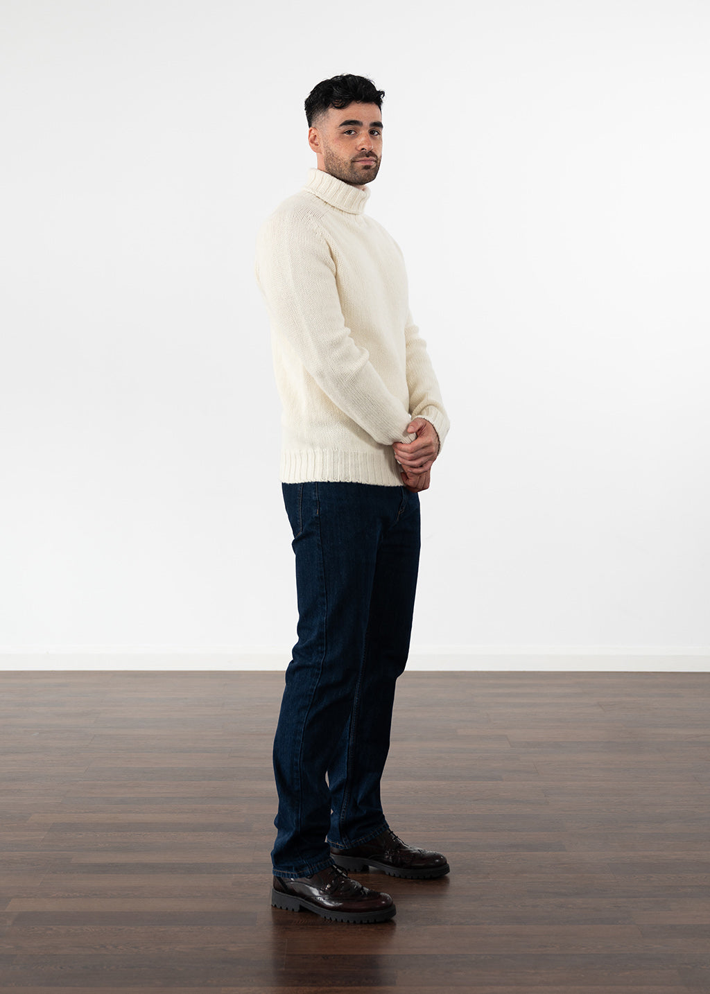 A man wearing a cream polo neck jumper, dark blue jeans, and dark burgundy leather shoes, standing in profile against a white wall and wooden floor.