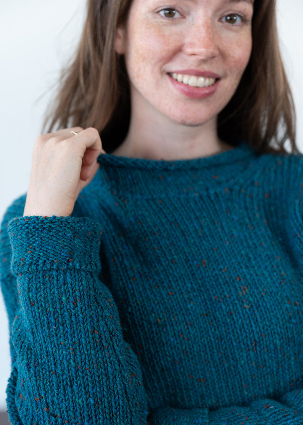 A smiling woman wearing a teal blue chunky-knit jumper with orange flecks, shown in a close-up portrait against a plain light background.
