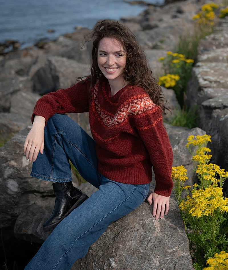 Girl wearing a russet and orange fair isle textured jumper whilst sitting on rocks by the sea, surrounded by yellow flowers