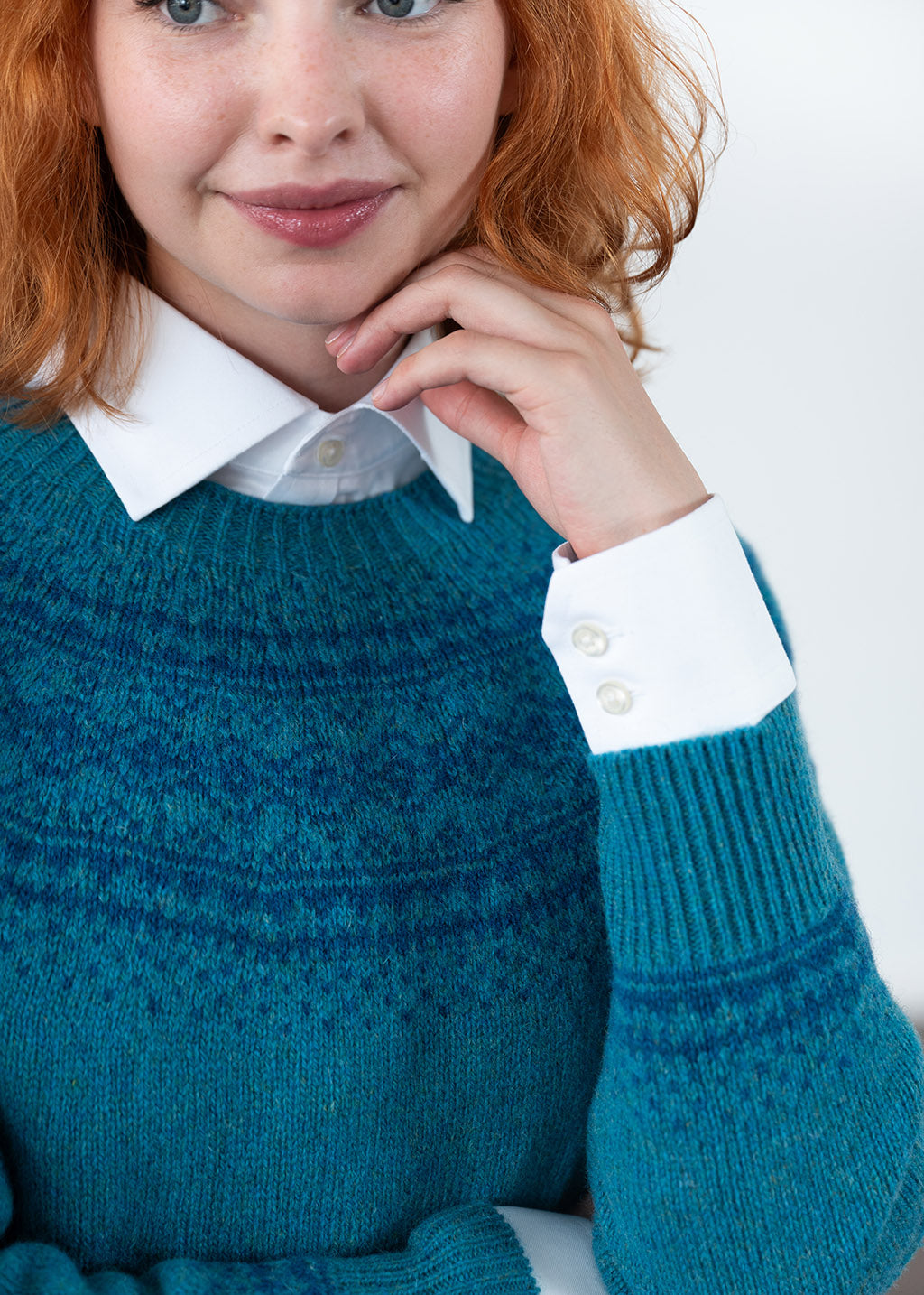 A close-up of a woman wearing a teal blue fair isle knit jumper over a white collared shirt, with her hand resting near her chin against a plain light background.