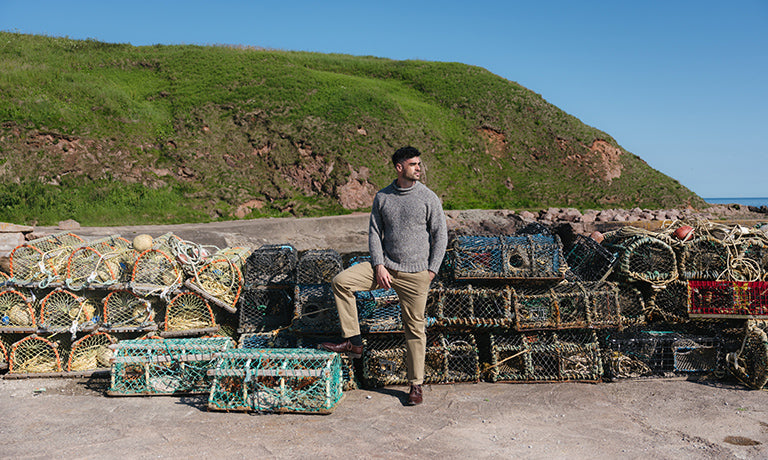 A man wearing a grey knit roll neck jumper and khaki trousers stands outdoors in front of stacked lobster pots on a rocky shore, with a green grassy hill and blue sky behind him.