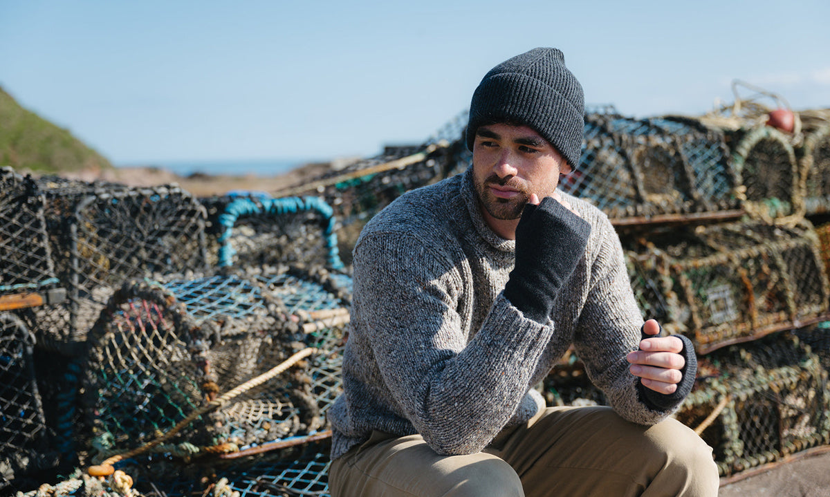 A man wearing a dark grey knit beanie, light grey knit jumper, and dark grey knit gloves sits in front of stacked lobster traps on a rocky beach.