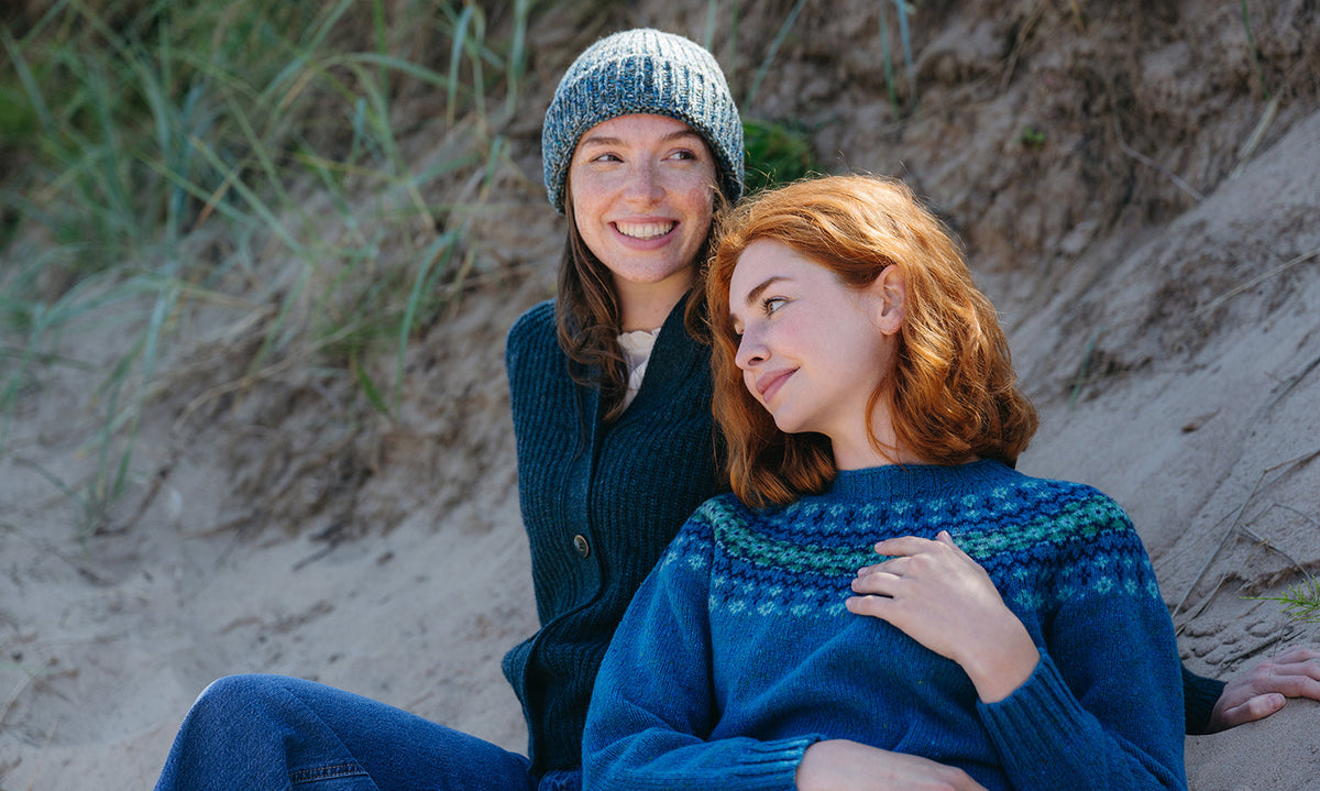 Two women sitting on a sandy dune outdoors, wearing cosy knit sweaters and a beanie. The woman in front wears a teal fair isle jumper, and the woman behind wears a dark teal cardigan over a light top.