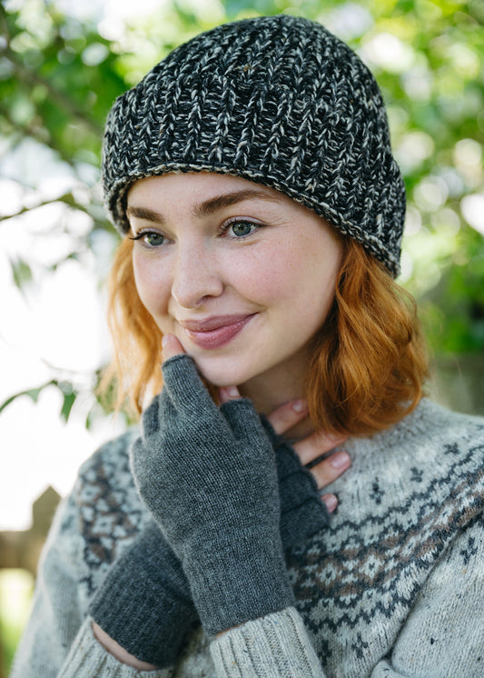 A woman outdoors wearing a charcoal and white marled knit beanie, mid grey fingerless gloves, and a grey fair isle jumper, with soft-focus green foliage background.