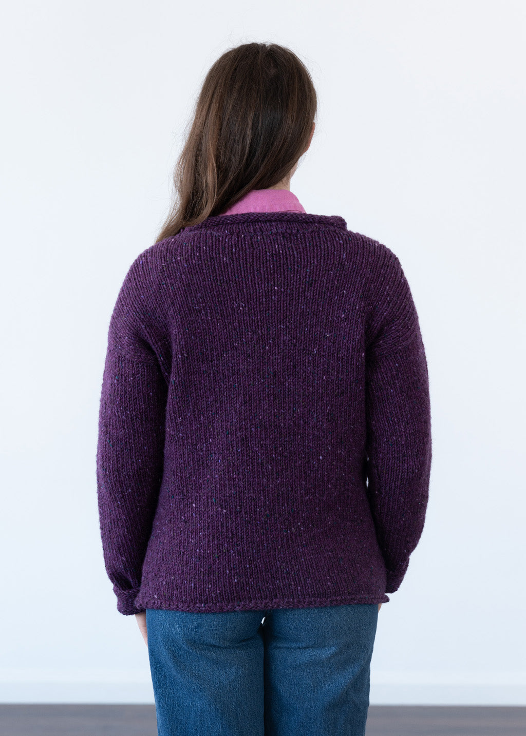 A woman viewed from behind wearing a textured, chunky knit purple jumper over a pink shirt and blue jeans, standing against a plain white wall.