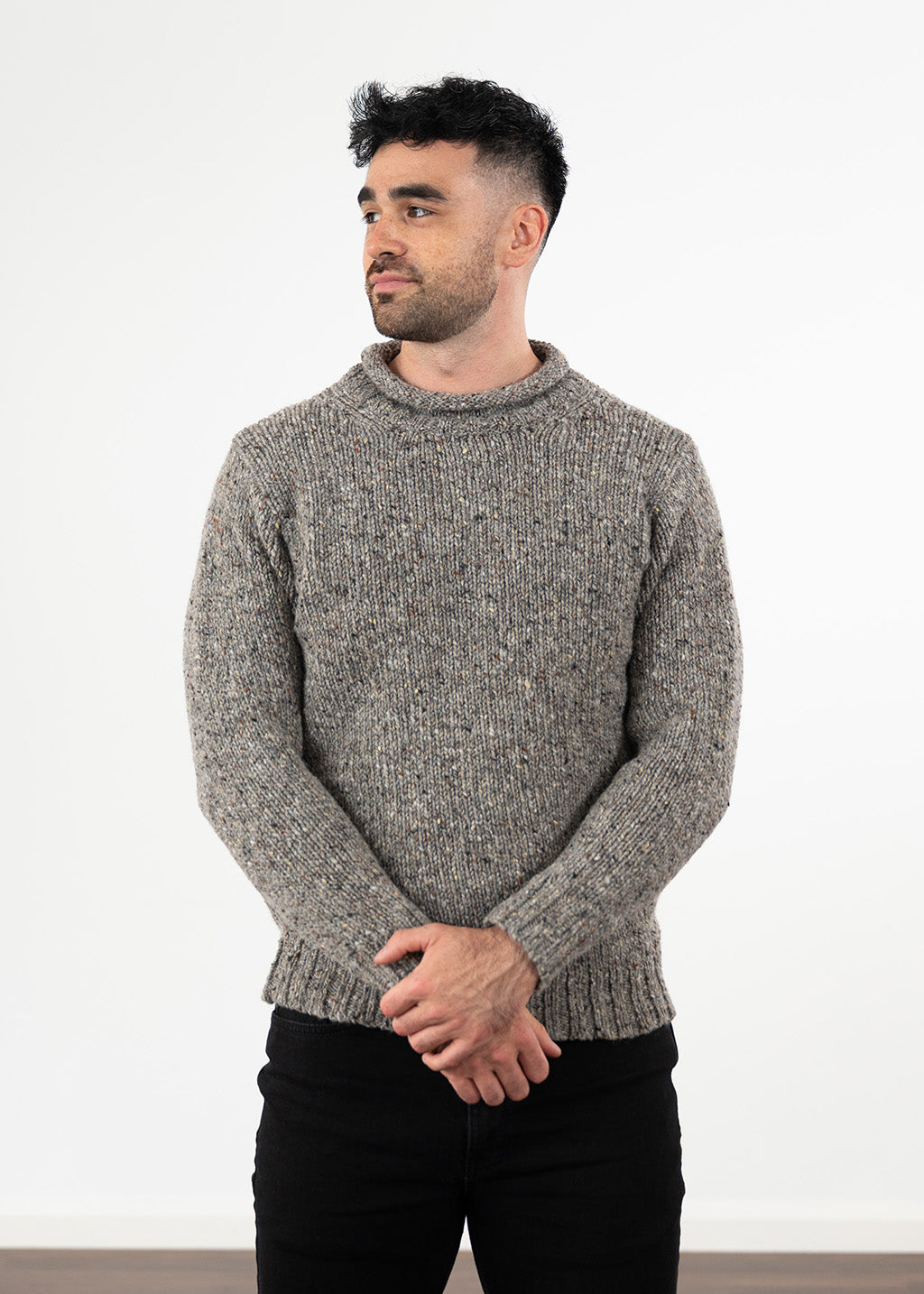 A man wearing a textured wool grey flecked knit jumper with a rolled crew neck, paired with black jeans, standing against a plain white wall.