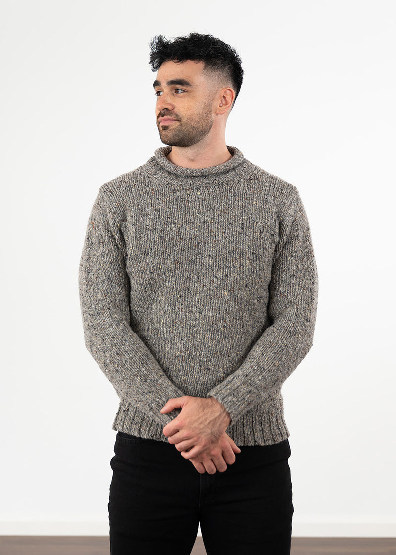 A man wearing a textured wool grey flecked knit jumper with a rolled crew neck, paired with black jeans, standing against a plain white wall.