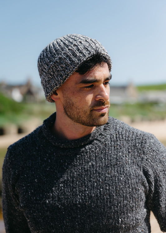 A man wears a mid grey marled chunky beanie hat with dark grey flecked jumper against an outdoor green blurred background.