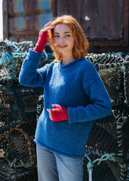 A woman with red hair wearing a chunky blue knit sweater and bright red wrust warmer fingerless gloves, posing in front of stacked lobster traps.