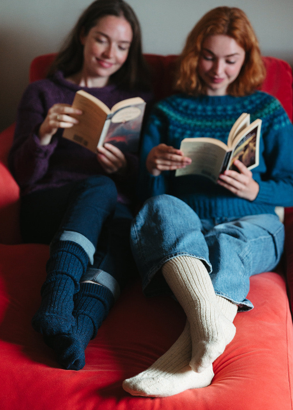 Two women lounging on a red settee whilst reading books. The focus is on their feet where they wear ribbed wool socks in navy and beige, pulled up to mid calf.