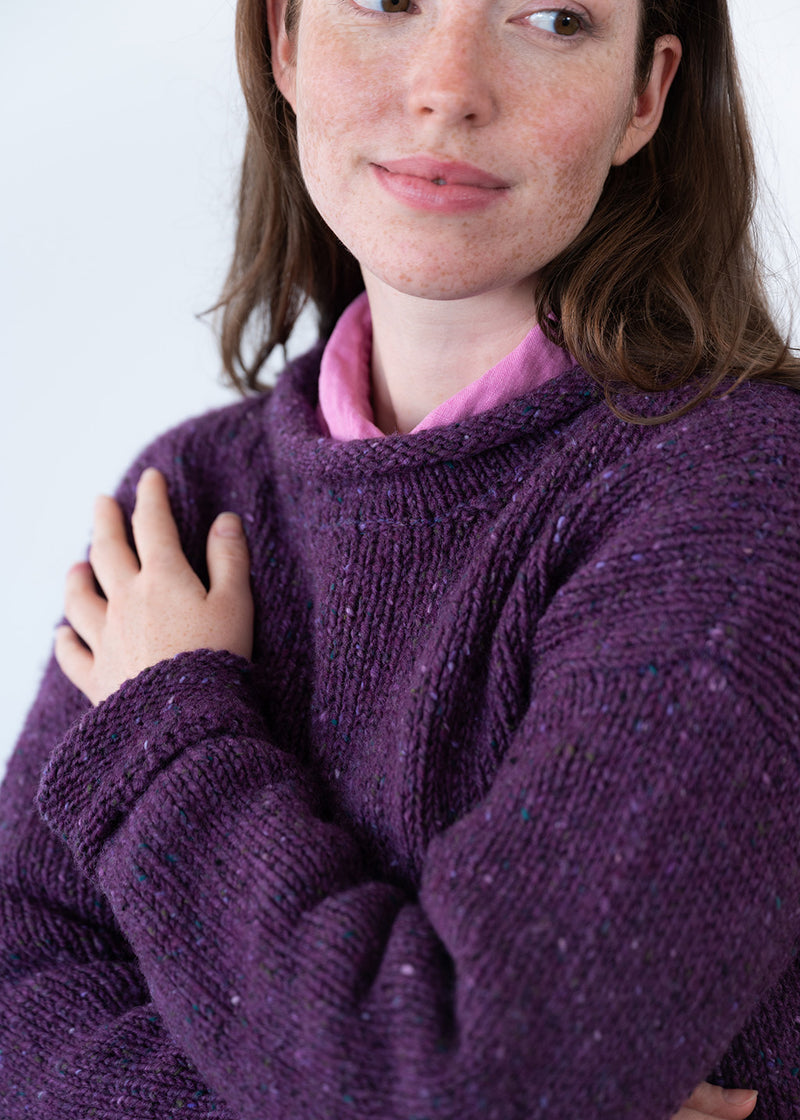 A close-up of a woman wearing a chunky knit purple jumper with subtle multicolor flecks, layered over a pink shirt, against a plain light background.