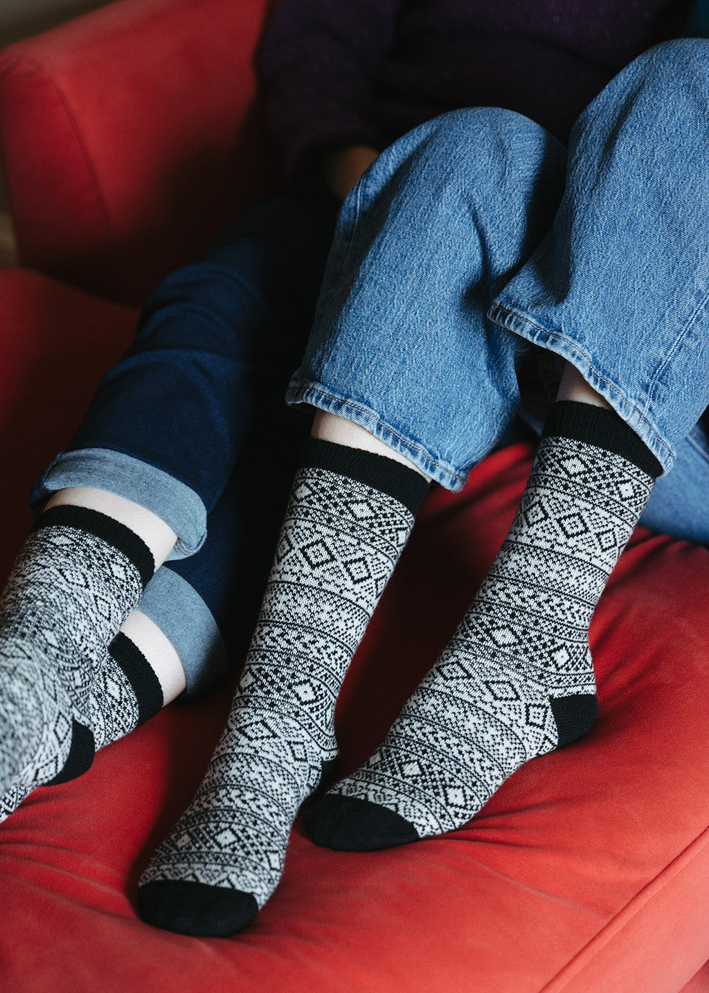 A close up of 2 people's legs lying on a red settee, both wearing fair isle patterned socks in light beige with black pattern and rib, heal and toes
