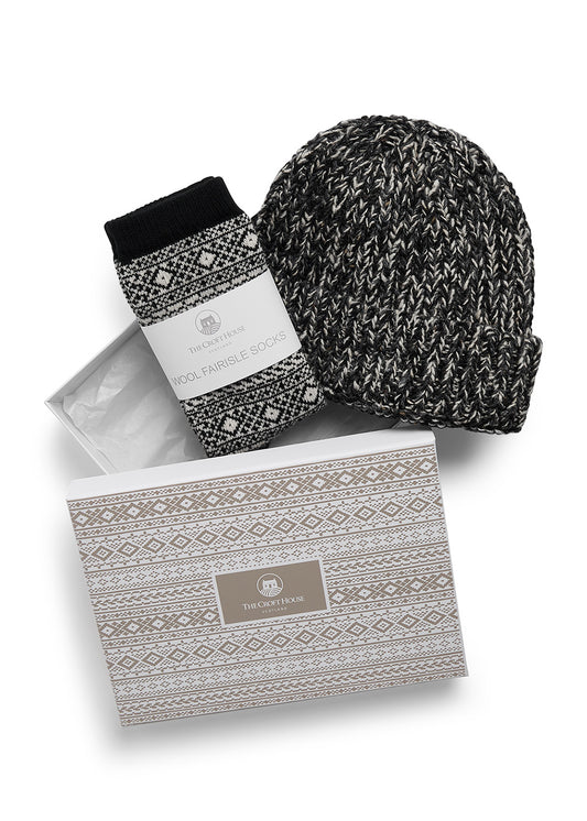 A pair of folded black and beige fair isle wool socks and co-ordinating marled wool beanie hat lying on top of a gift box with beige fair isle patterns and The Croft House logo on the lid, against a white background.