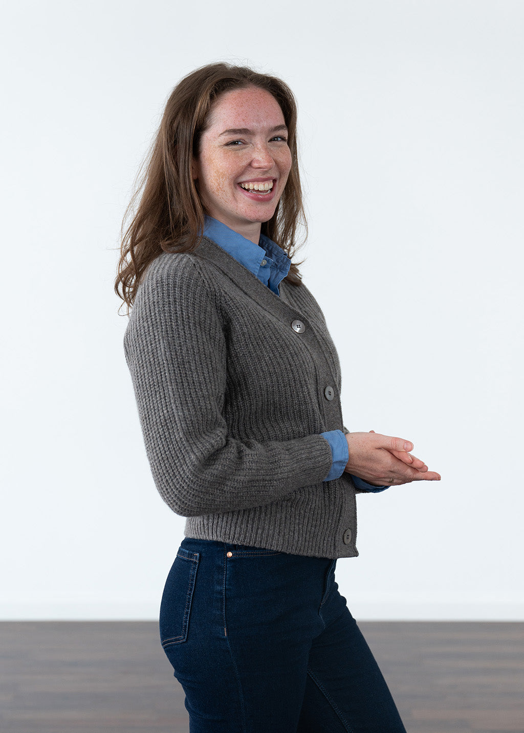 A woman wearing a brown ribbed cardigan over a blue collared shirt and dark blue jeans, standing against a plain white studio background.