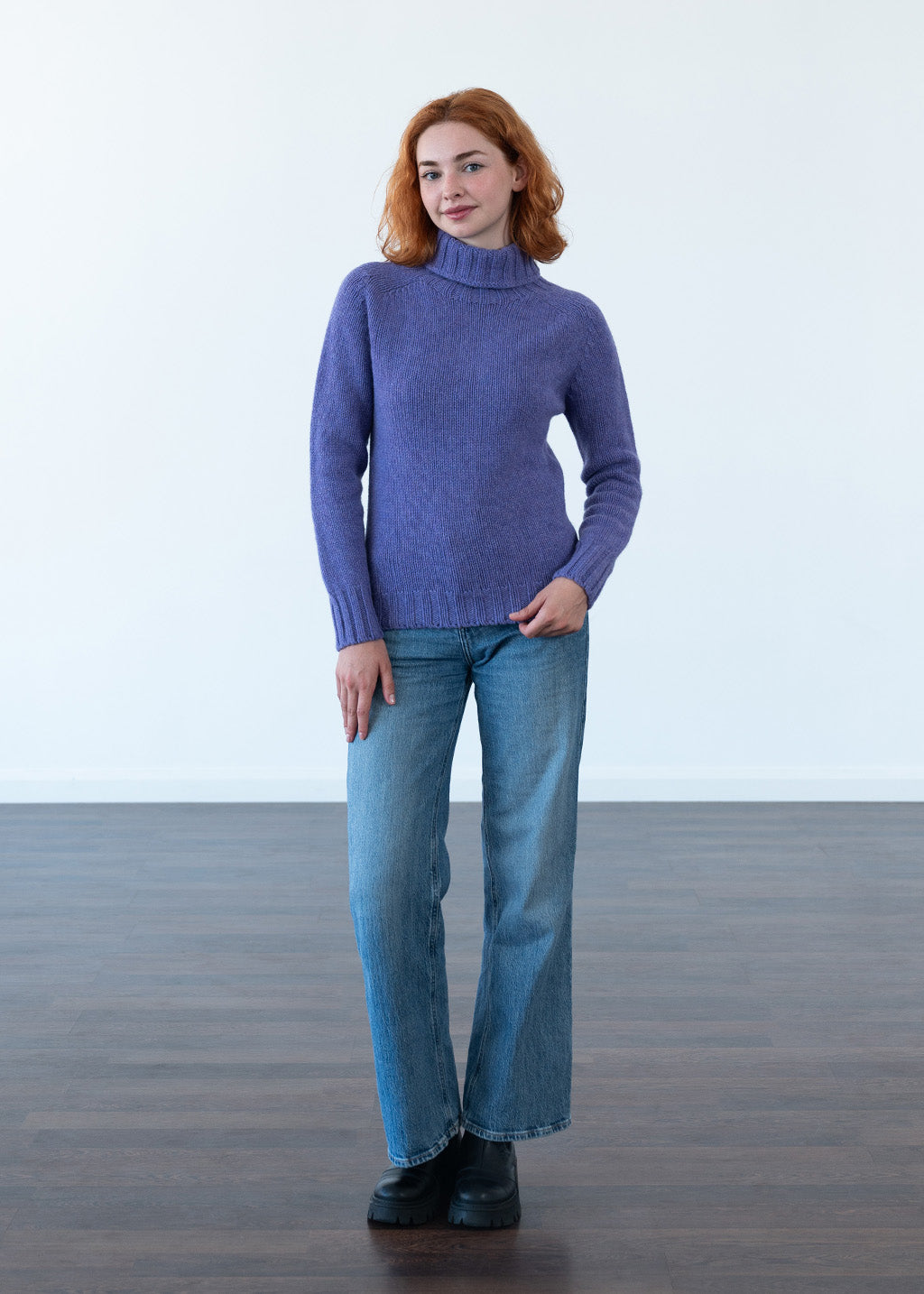 A woman with red hair stands in a studio wearing a purple polo neck jumper, blue jeans, and black boots against a white wall and dark wood floor.