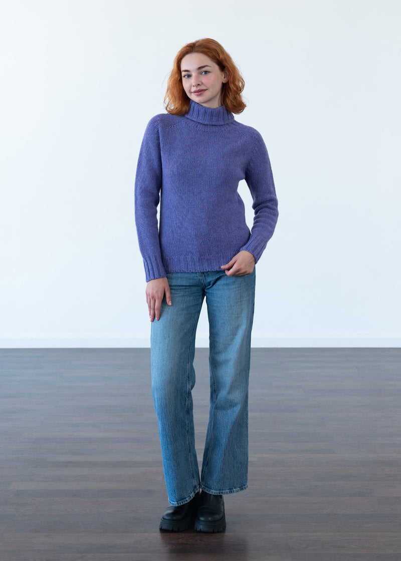 A woman with red hair stands in a studio wearing a purple polo neck jumper, blue jeans, and black boots against a white wall and dark wood floor.