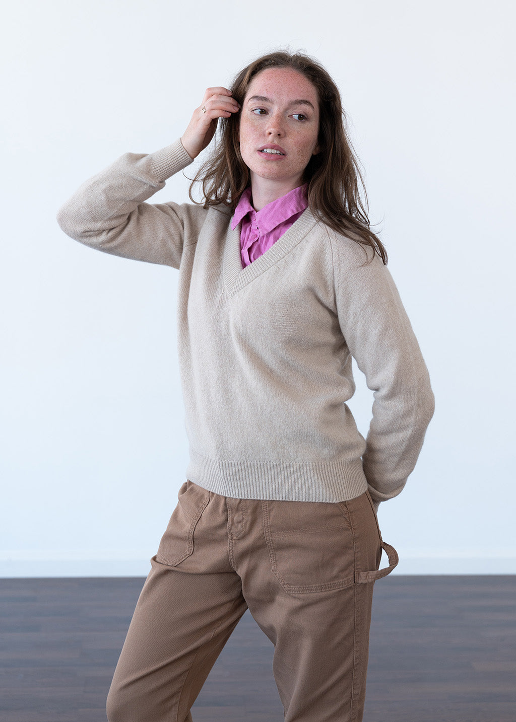 A woman modeling a beige wool V-neck sweater over a pink collared shirt and brown pants, standing against a plain white studio background.