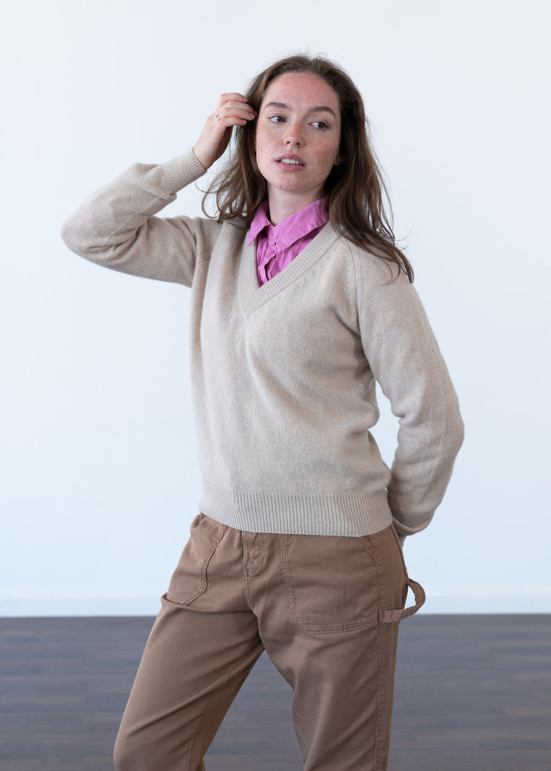 A woman modeling a beige wool V-neck sweater over a pink collared shirt and brown pants, standing against a plain white studio background.
