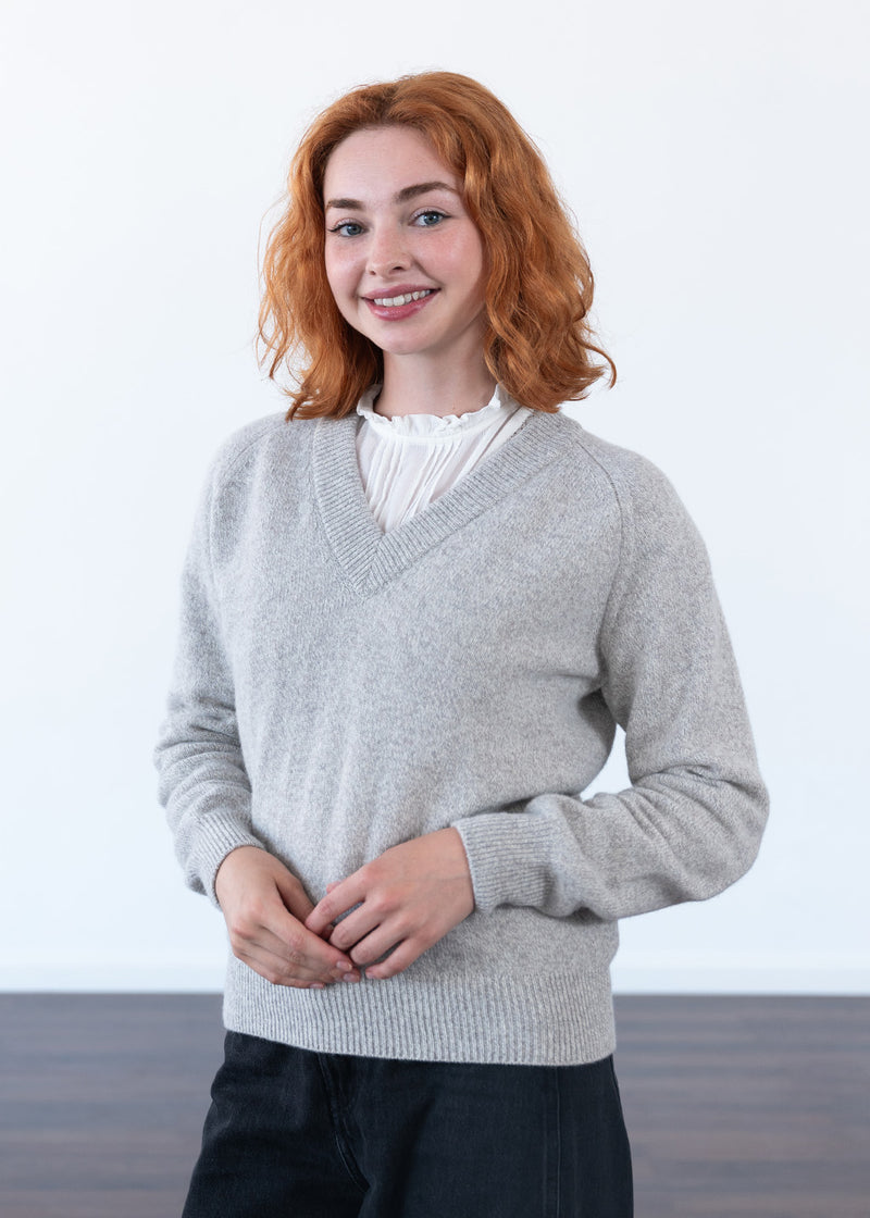 A woman with red hair wearing a light grey marl v-neck knit jumper over a white ruffled blouse and dark jeans, standing against a plain white wall.