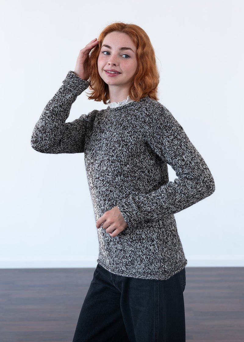 A woman with red hair wearing a marled ecru and black knit sweater over a white collar shirt, paired with dark jeans, standing against a white wall.