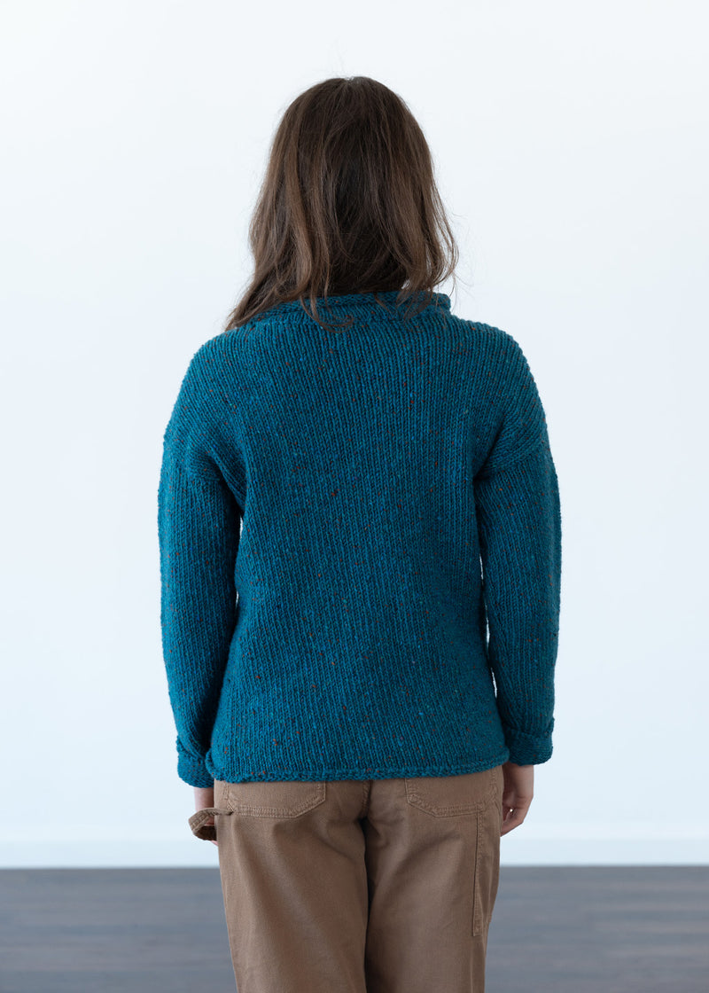 A person viewed from behind wearing a teal textured knit jumper and tan trousers against a clean white studio background.
