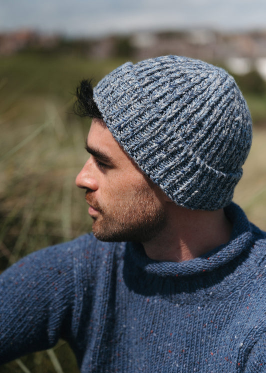 A man wearing a chunky wool beanie hat in shades of light denim blue and grey, in a coastal setting.