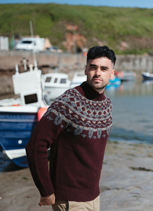 A man wearing a dark burgundy fair isle jumper with gray yoke pattern stands on a rocky shoreline with boats and a green hill in the background.