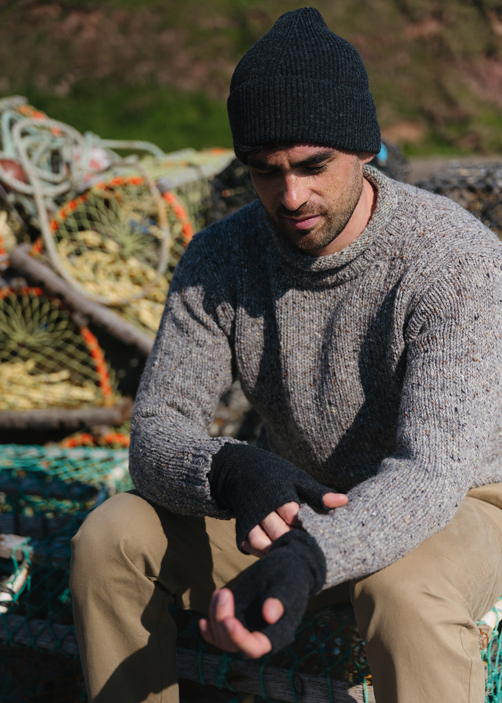 A man wearing a charcoal grey knit beanie, a light grey donegal knit jumper, and tan chinos sits outdoors on a dock, adjusting a charcoal grey fingerless glove on his hand. Fishing creels are visible in the background.
