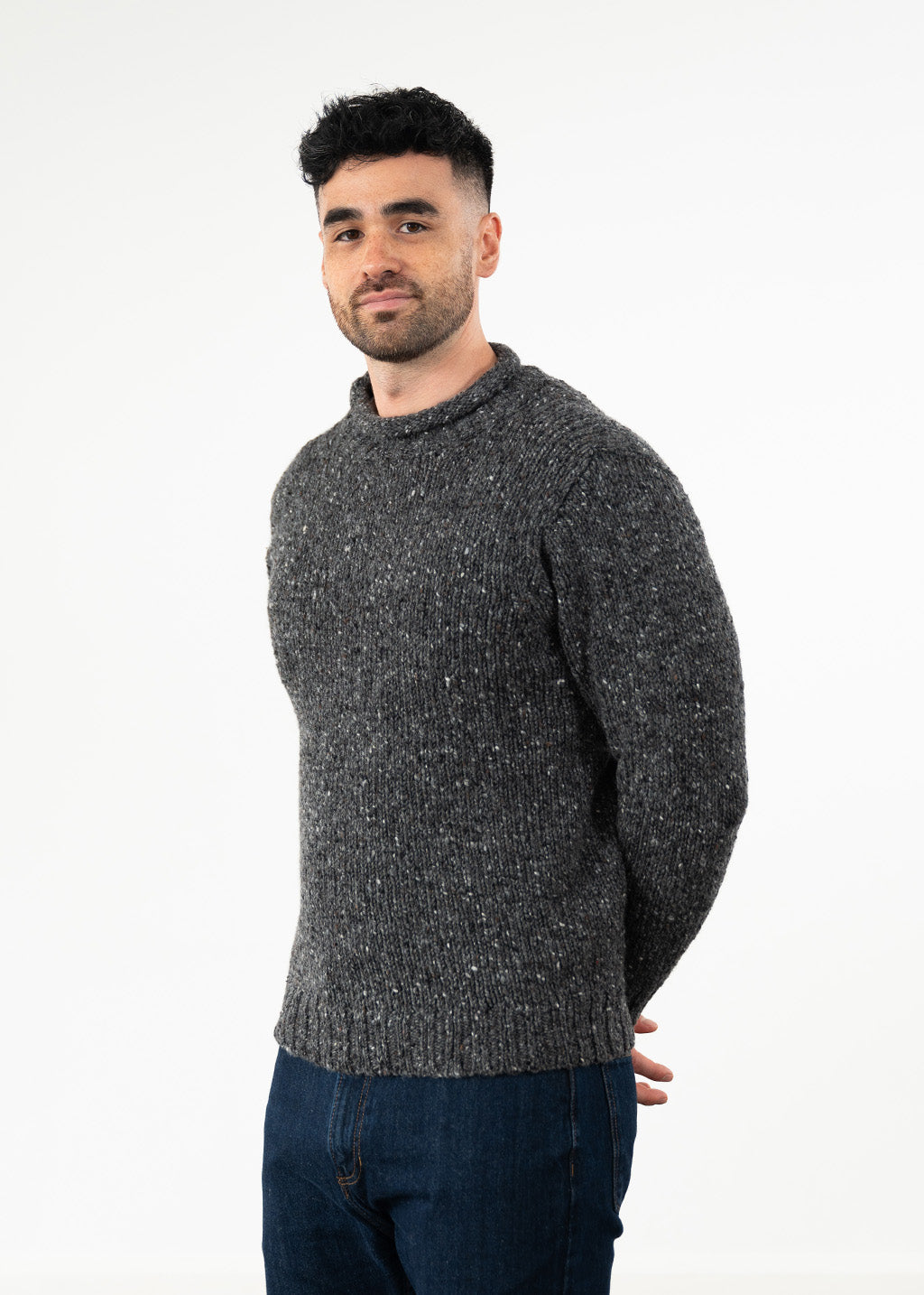 A man wearing a dark gray donegal wool flecked knit sweater and blue jeans, standing against a plain white studio background.