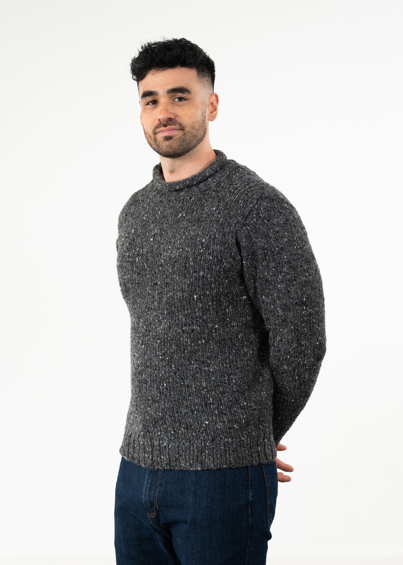 A man wearing a dark gray donegal wool flecked knit sweater and blue jeans, standing against a plain white studio background.