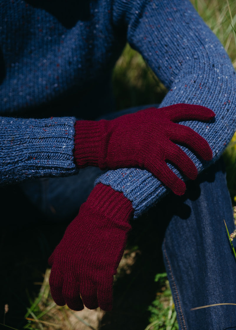 A close-up of a person wearing a blue knit jumper and blue jeans, with one hand raised to show a burgundy knit glove. The background is a blurred outdoor setting with grass and sunlight.