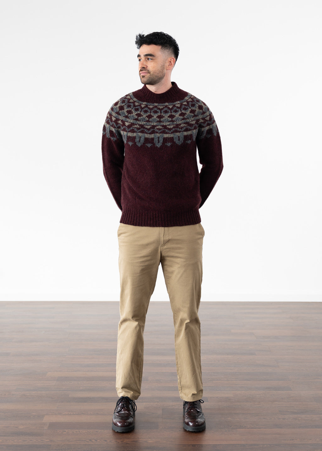 A man wearing a burgundy Fair Isle knit sweater with beige trousers and dark burgundy leather shoes, standing against a white studio background with a wooden floor.