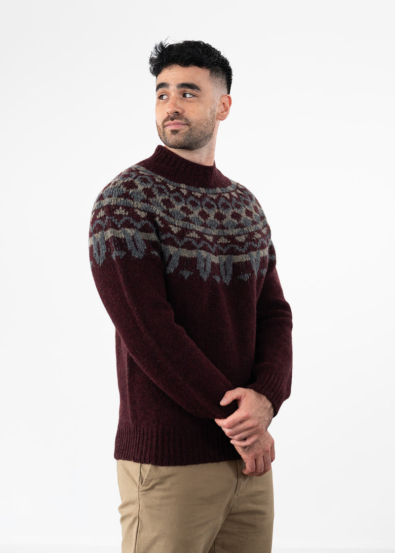A man wearing a burgundy knit sweater with a grey and beige Fair Isle yoke pattern, paired with khaki trousers, standing against a plain white studio background.