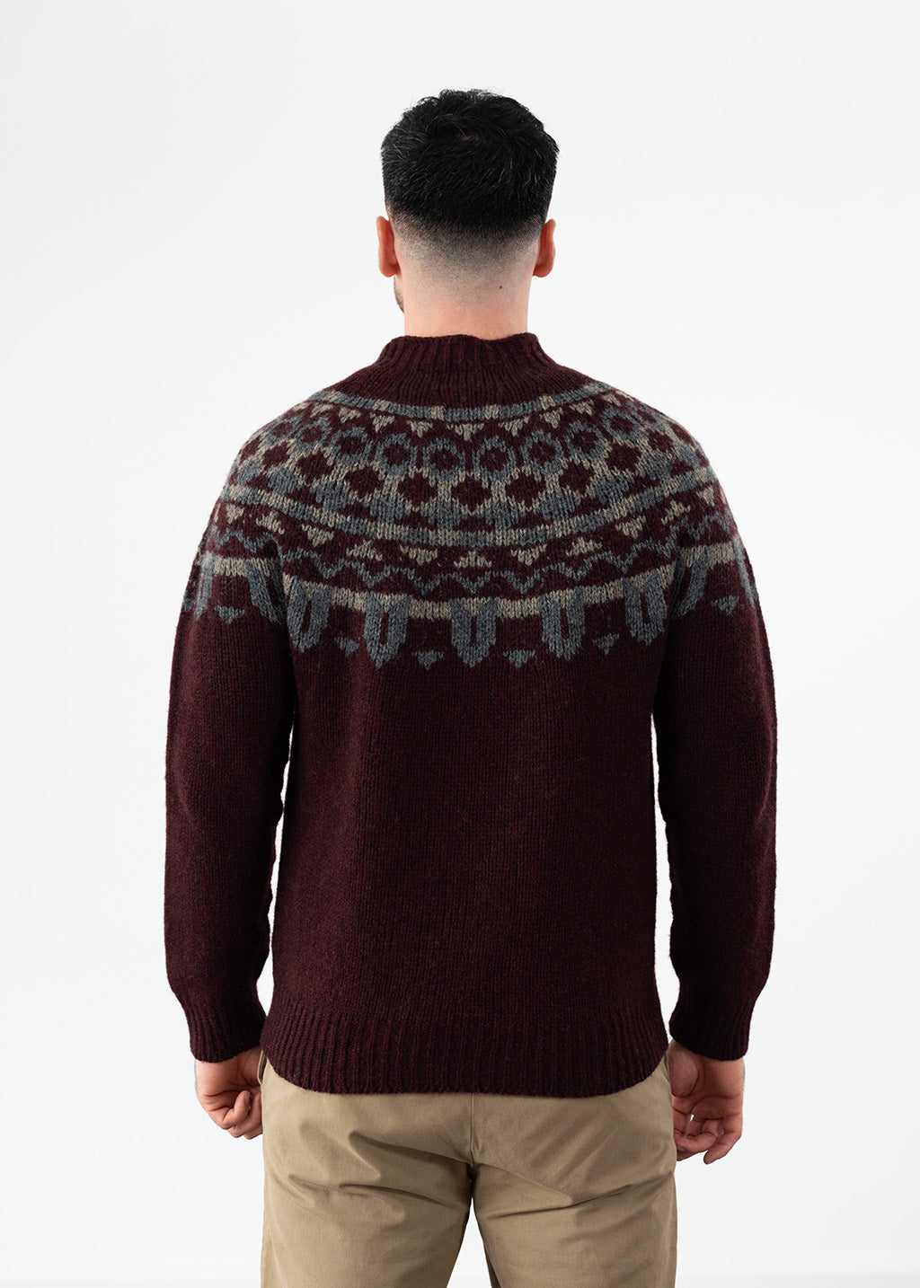 A man viewed from behind wearing a dark burgundy knit sweater with a gray and beige Fair Isle yoke pattern, paired with khaki pants, against a plain white studio background.