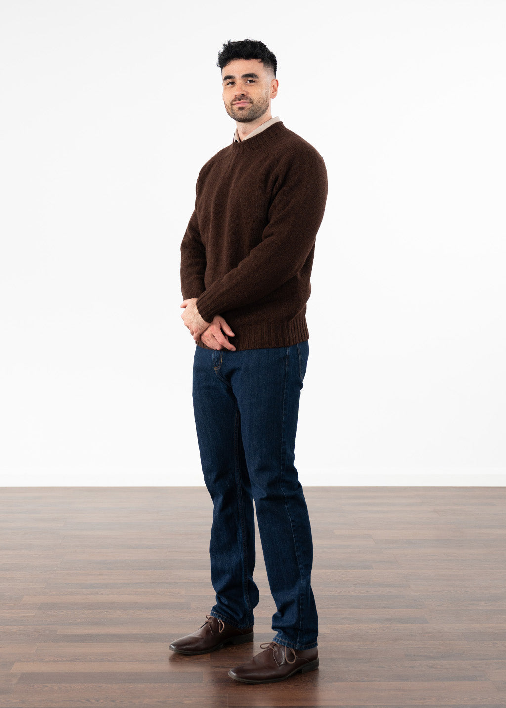 A man wearing a dark coffee brown knit crew neck jumper, dark blue jeans, and brown leather shoes, standing against a white studio background with a wooden floor.