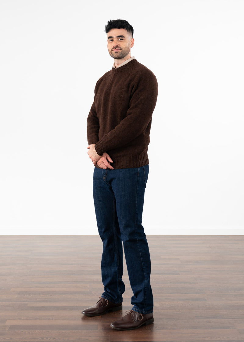A man wearing a dark coffee brown knit crew neck jumper, dark blue jeans, and brown leather shoes, standing against a white studio background with a wooden floor.
