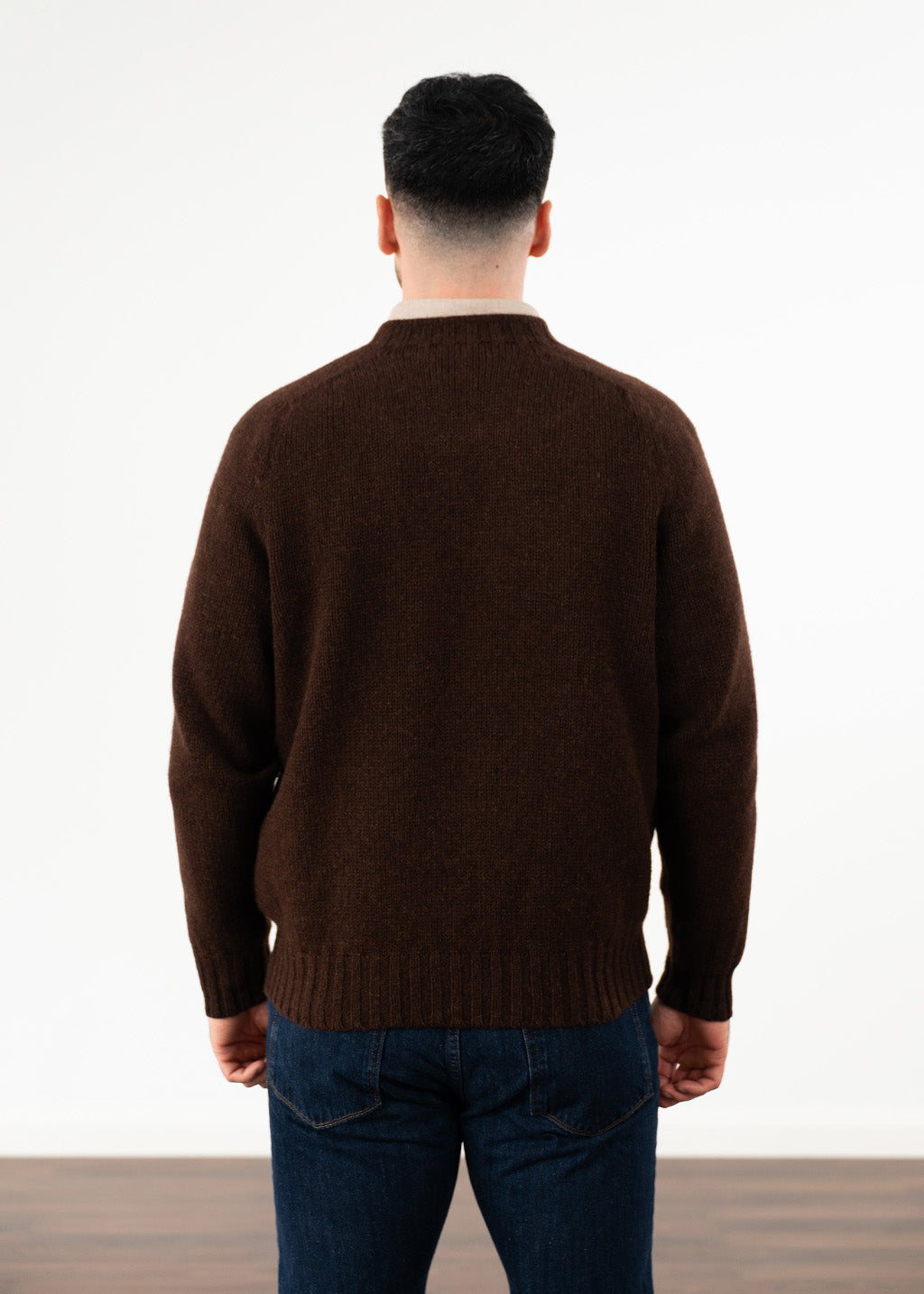 A man viewed from behind wearing a dark brown knit jumper and blue jeans, standing against a plain white studio background.