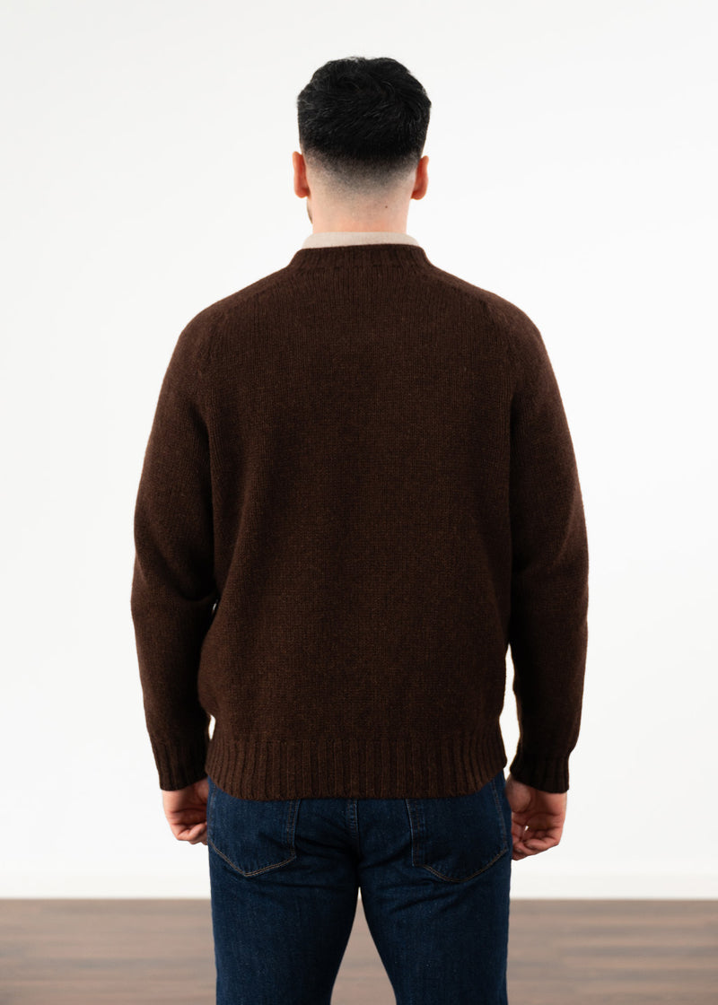 A man viewed from behind wearing a dark brown knit jumper and blue jeans, standing against a plain white studio background.