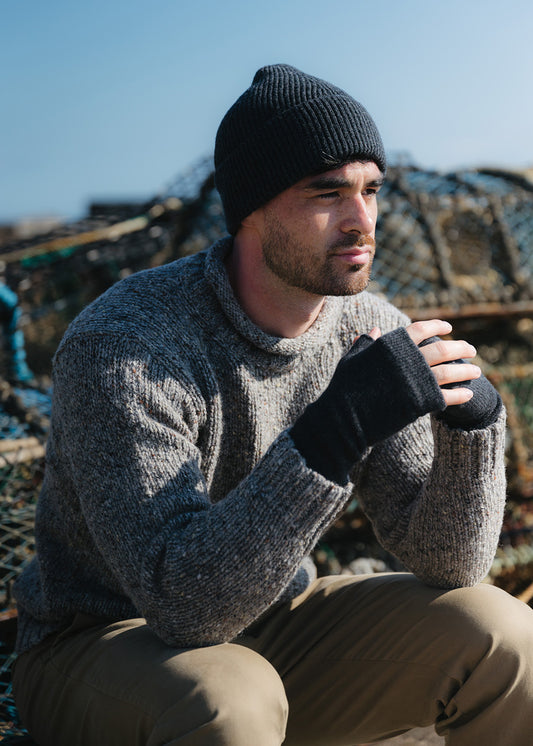 A man wearing a charcoal grey knit ribbed beanie, a grey flecked knit sweater, and charcoal grey fingerless wrist warmer gloves, sitting outdoors in front of fishing creels under a clear blue sky.