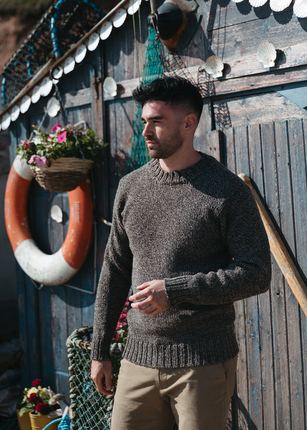 A man wearing a brown marl knit jumper and khaki trousers, standing in front of a rustic wooden wall decorated with nautical items like a life preserver, fishing nets, and seashells.