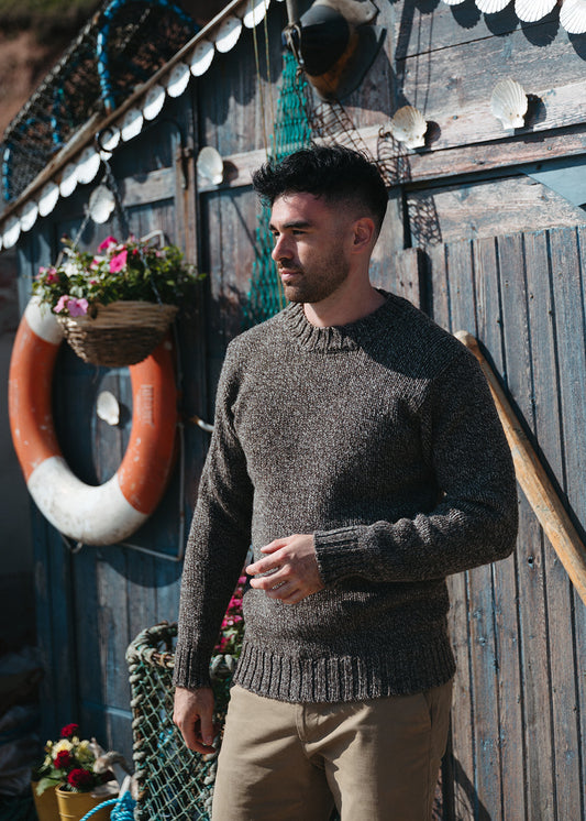 A man wearing a brown marl knit jumper and khaki trousers, standing in front of a rustic wooden wall decorated with nautical items like a life preserver, fishing nets, and seashells.