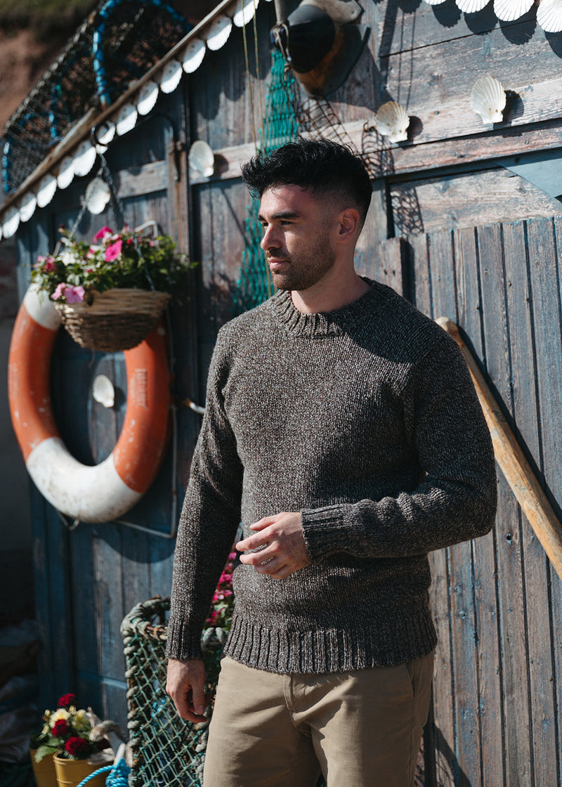 A man wearing a brown marl knit jumper and khaki trousers, standing in front of a rustic wooden wall decorated with nautical items like a life preserver, fishing nets, and seashells.