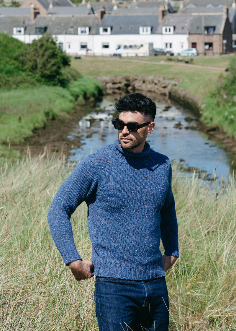A man wearing a textured blue knit wool jumper and dark sunglasses stands outdoors in a grassy field next to a small stream, with houses in the background.