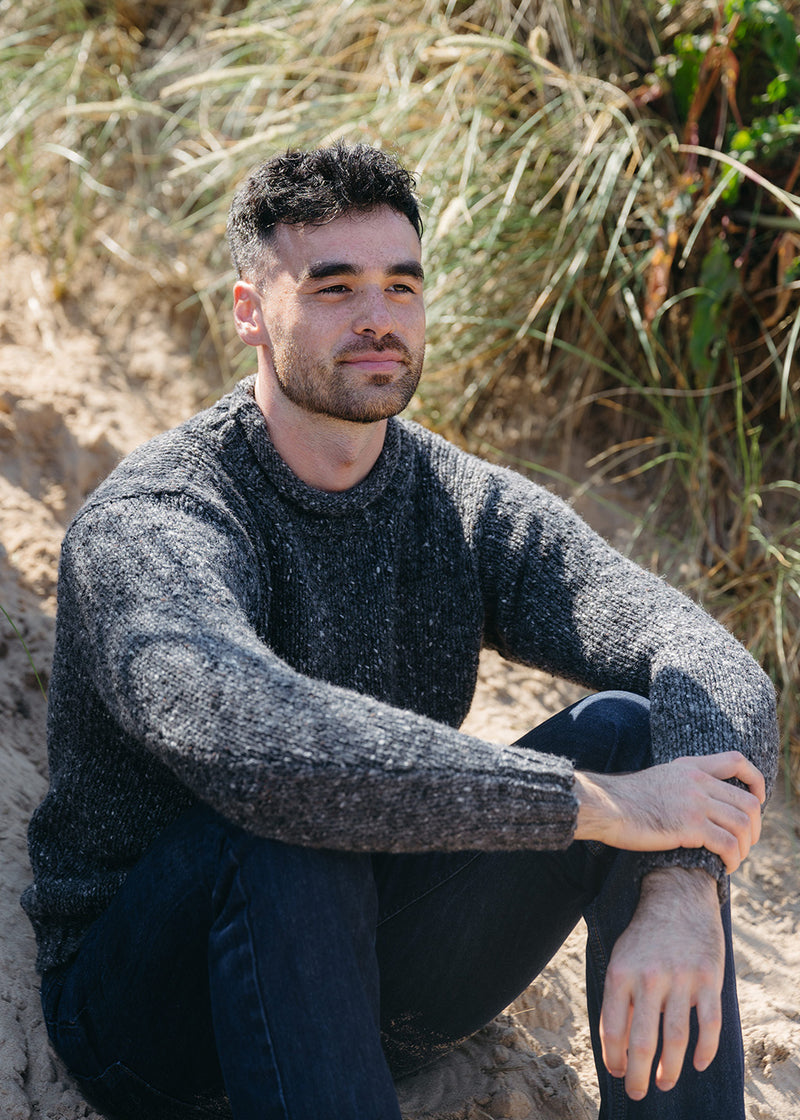 A man wearing a dark gray donegal knit jumper and dark jeans sitting on sandy dunes with beach grass in the background.