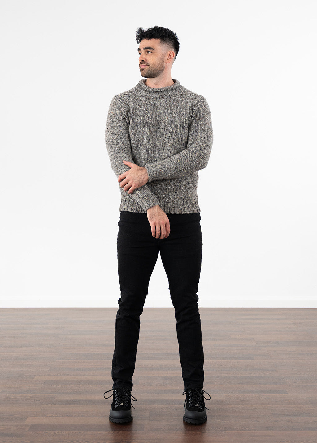 A man wearing a grey textured knit roll neck jumper, black slim-fit jeans, and black lace-up boots, standing against a white studio background with a wooden floor.