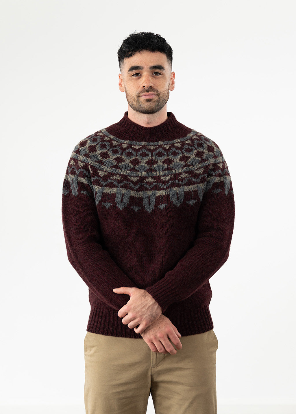 A man wearing a dark burgundy knit jumper with a grey and beige Fair Isle yoke pattern, paired with beige chinos, standing against a plain white studio background.