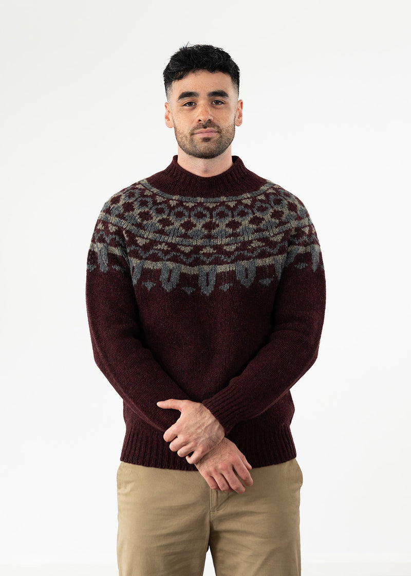 A man wearing a dark burgundy knit jumper with a grey and beige Fair Isle yoke pattern, paired with beige chinos, standing against a plain white studio background.