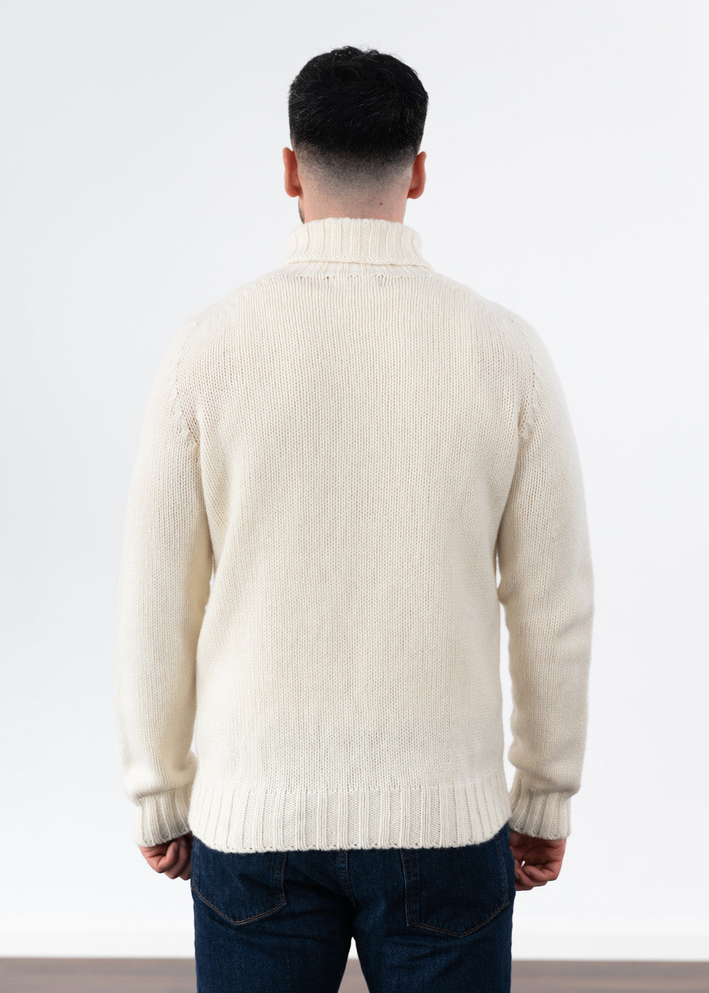 A man viewed from behind wearing a cream polo neck jumper and dark blue jeans, standing against a plain white studio background.
